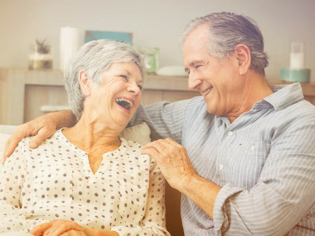 An elderly man and woman sitting on a couch, smiling and laughing together. The man has gray hair, wearing a striped shirt, and his arm around the woman, who has short gray hair and a patterned blouse. Cozy indoor setting with soft lighting.