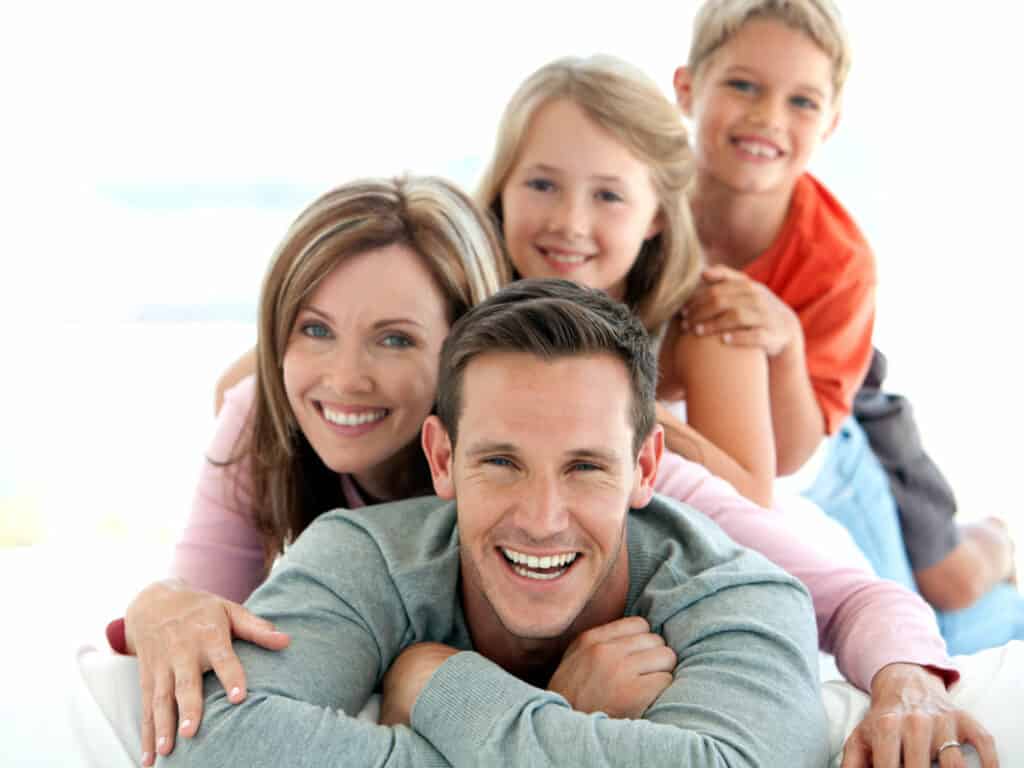 A happy family of four lying on a bed, stacked in a playful manner. A man in front, smiling, with a woman and two children behind him, all smiling at the camera. The background is bright and airy.