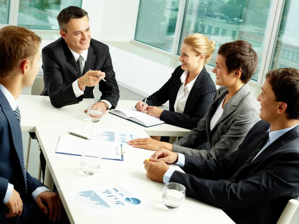 Five business professionals in a meeting room, sitting around a white table. They are engaged in discussion, with documents, charts, and glasses of water on the table. The room has large windows providing natural light.