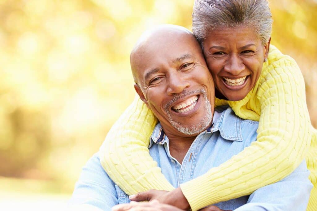 An older couple laughing together outdoors. The woman, wearing a yellow sweater, lovingly embraces the man from behind, who is in a denim shirt. The background is a soft, out-of-focus golden color, suggesting a sunny day.