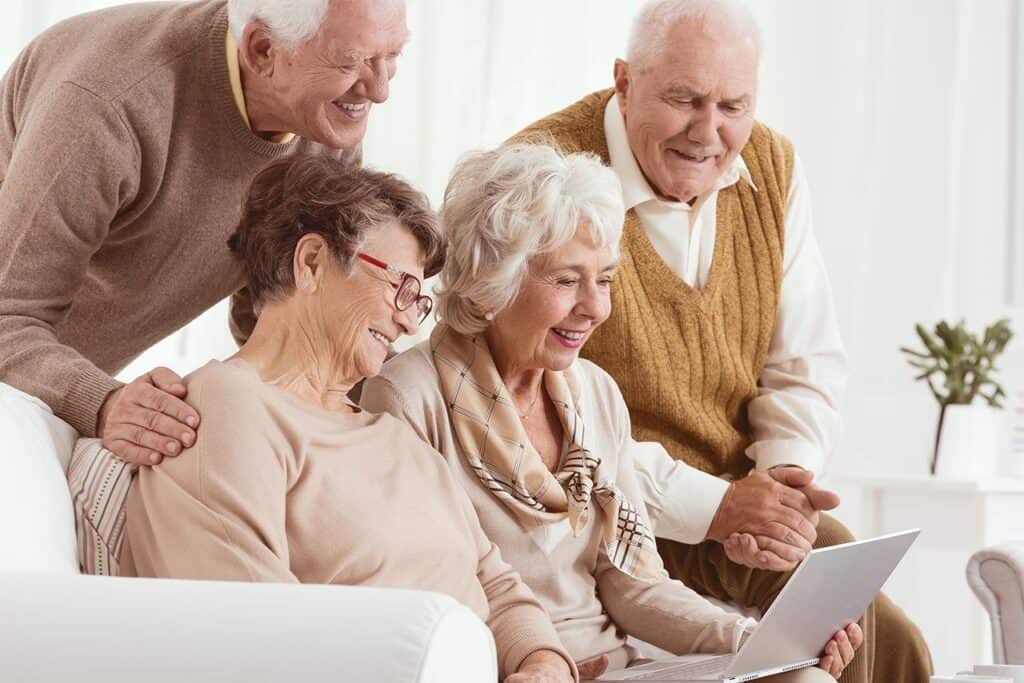 Four elderly people are gathered around a laptop on a cozy white sofa. Two men stand behind two seated women. They are smiling and appear engaged with the screen, suggesting a relaxed, happy interaction.
