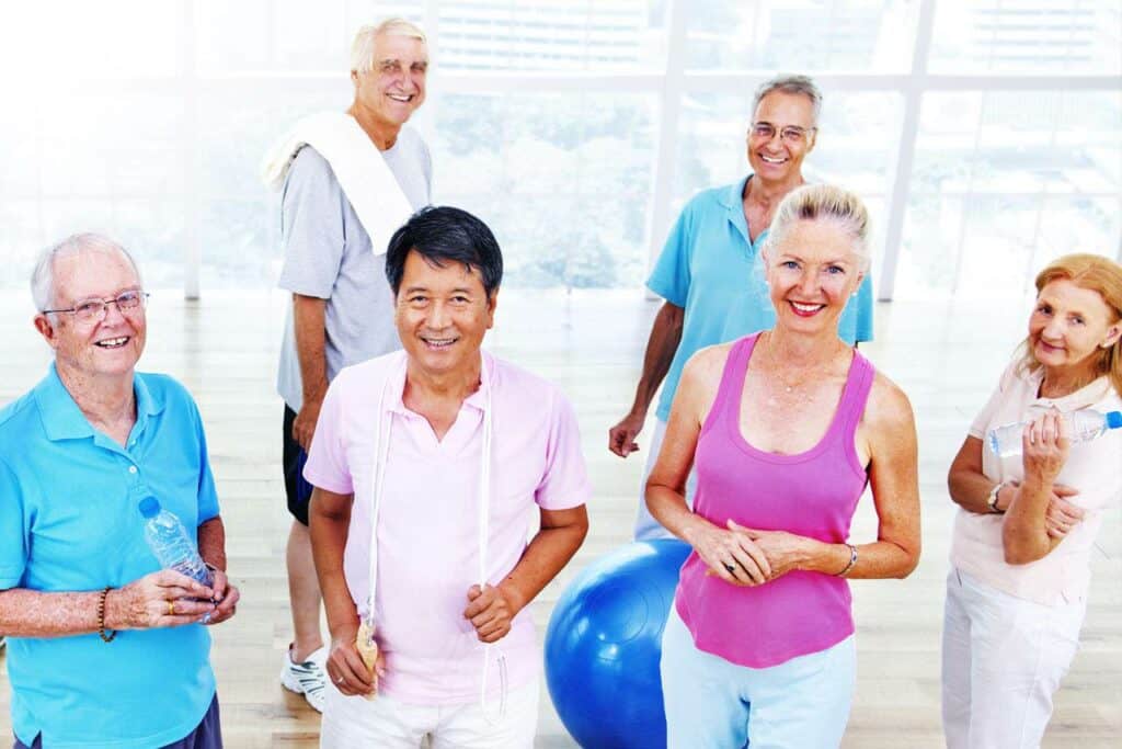 A group of six older adults in fitness attire smile and pose in a bright, spacious gym. They hold water bottles and a towel, with an exercise ball visible in the background. Large windows reveal an urban scene outside.