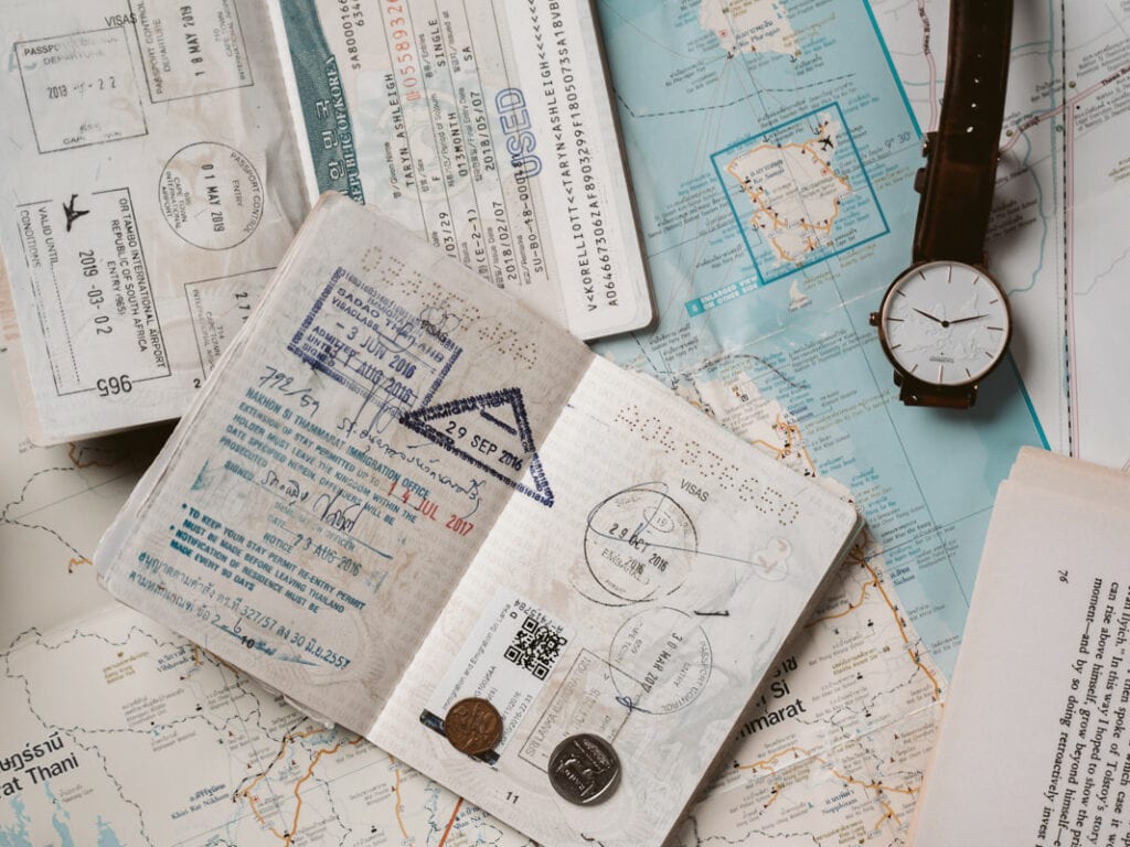Open passport with stamps on top of maps, surrounded by travel documents, coins, and a wristwatch. Documents show various dates and stamps, suggesting international travel. Maps and items indicate planning or reminiscing about past journeys.