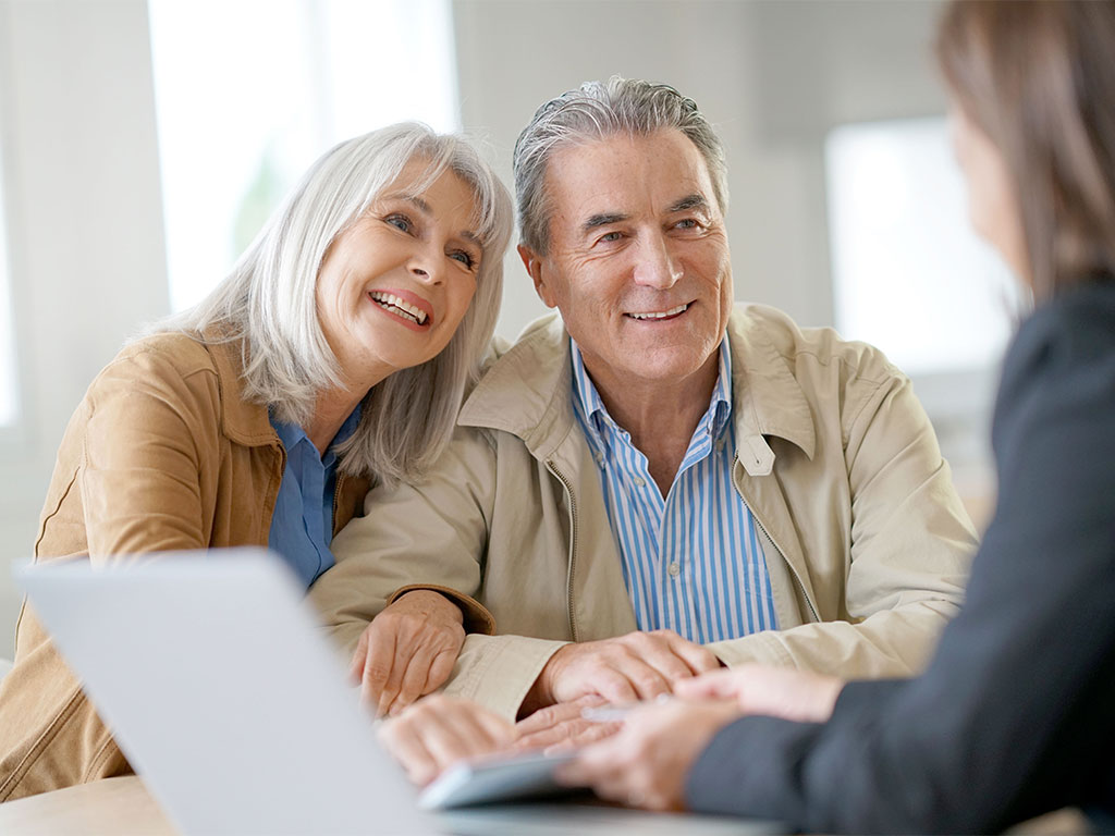 An older couple sits at a desk, smiling and listening to a professional woman across from them. papers and a laptop are on the table, suggesting a meeting or consultation in a bright, modern office.