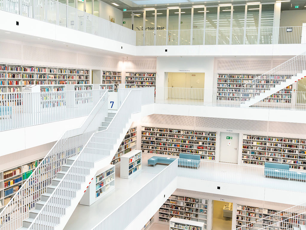 A modern, multi level library with white walls and railings, open staircases, and rows of bookshelves filled with books. the space is bright and airy with natural light from large windows.